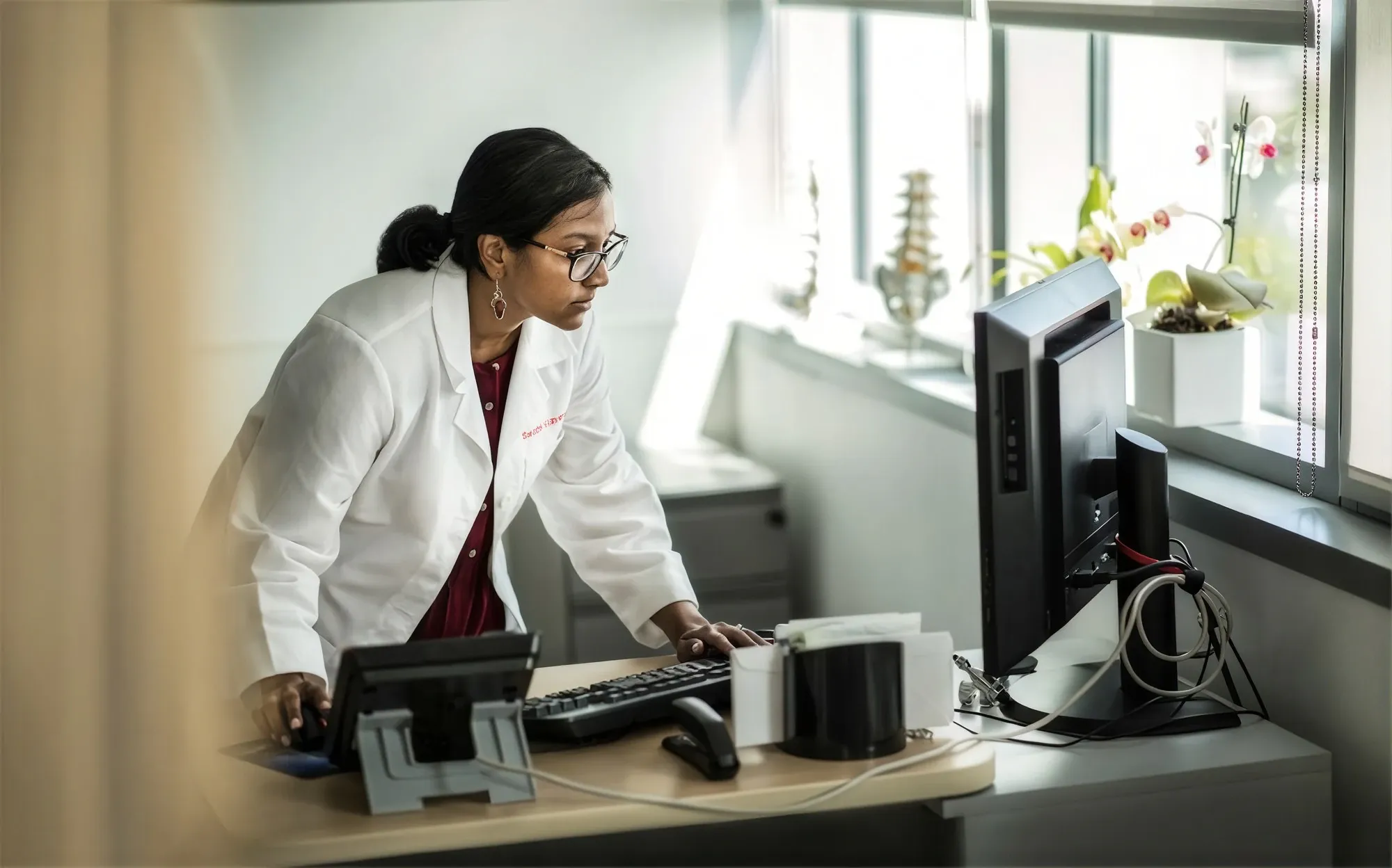 A doctor in a white coat working at a computer by a clinic window
