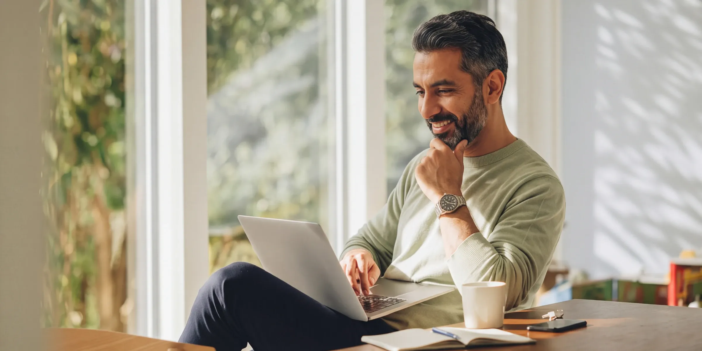 A smiling man in a green sweater working on a laptop beside a sunlit window