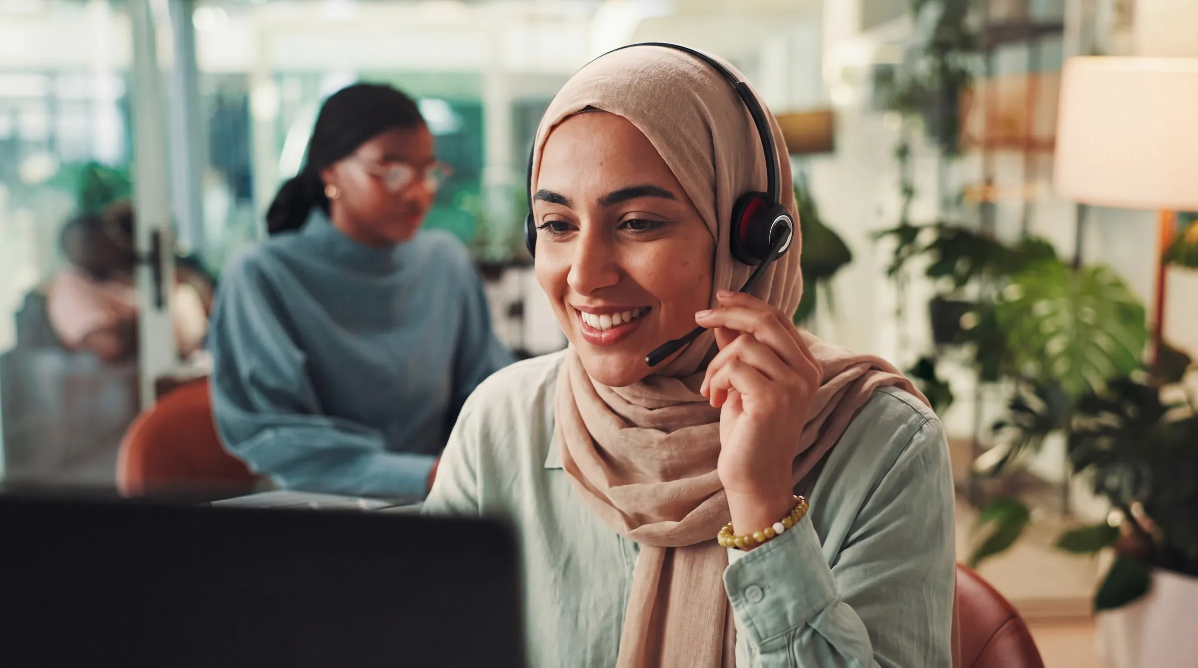 A smiling contact-center agent in a headscarf adjusting her headset at her desk